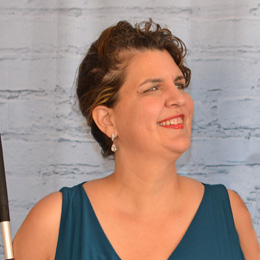 Amy Porterfield smiles warmly while holding a white cane. She has short, curly brown hair and pearl drop earrings. She wears a sleeveless teal dress.