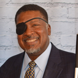 President Donald Porterfield wears a friendly smile while holding a cane. He has short, dark hair and a neatly trimmed beard. He is wearing a black eye patch over his right eye, dark suit jacket, a light blue dress shirt, and a blue and yellow patterned tie.